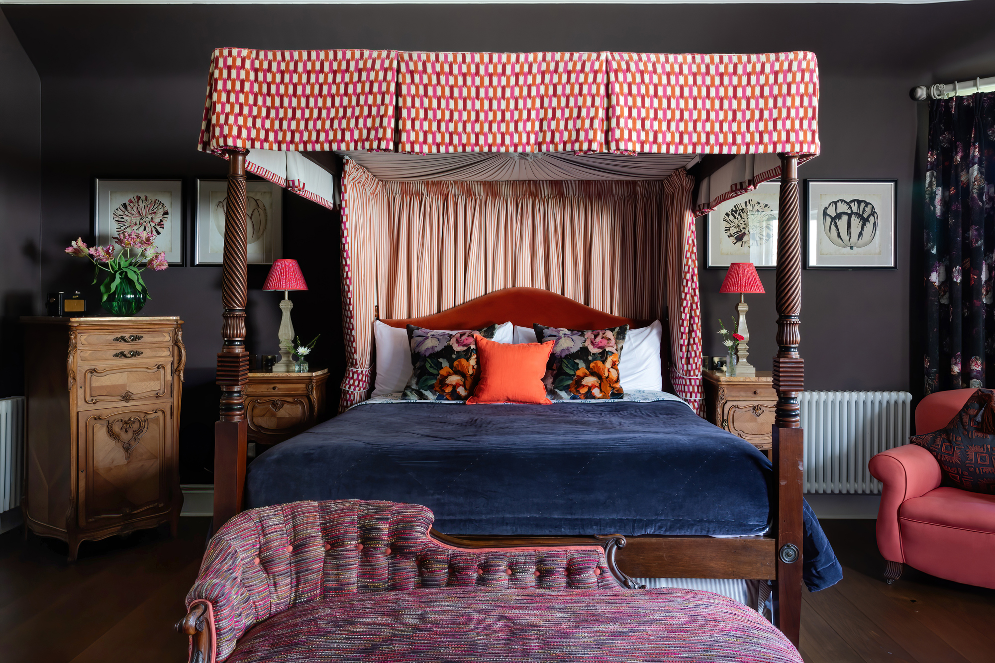 A richly decorated bedroom at the Cromlix Hotel featuring a grand four-poster bed with patterned drapes and canopy, a dark blue velvet bedspread, floral and orange pillows, bedside tables with red lamps, and a patterned chaise lounge in the foreground.